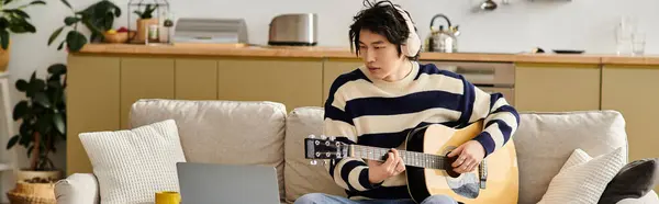A handsome young man practices playing his acoustic guitar while seated on a cozy couch, banner — Stock Photo