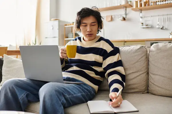 Focalizzato giovane asiatico uomo studi online dal comfort della sua casa accogliente sorseggiando una bevanda. — Foto stock