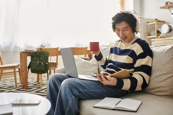 A cheerful student on a couch in a bright room learns online while sipping coffee. — Stock Photo