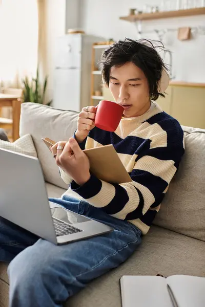 Young man enjoys his studies at home, sipping coffee while using his laptop and taking notes. — Stock Photo