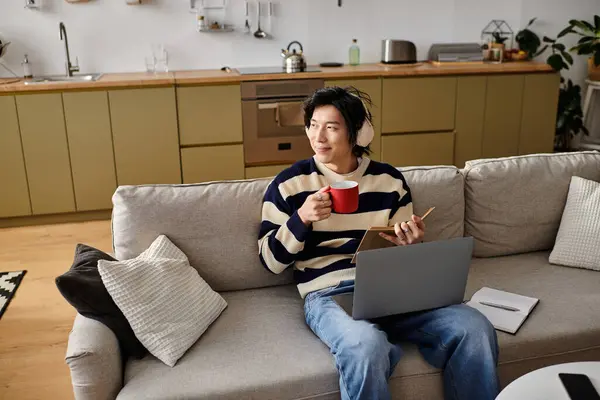 A handsome young man studies from home, enjoying a warm drink and focused on his laptop. — Stock Photo