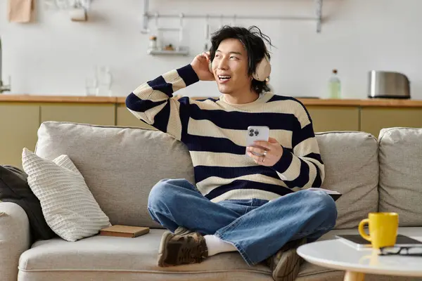 Relajándose en casa, un joven escucha música mientras usa su teléfono y sonríe felizmente. - foto de stock