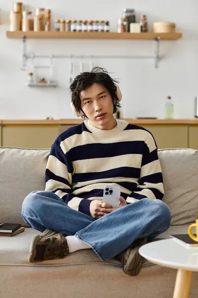 Un joven guapo se sienta casualmente en un sofá en casa, sosteniendo su teléfono mientras se relaja. - foto de stock