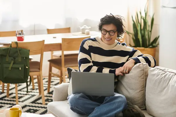 Handsome young asian man smiles while studying on his laptop from a cozy corner of his home. — Stock Photo