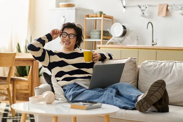 A young man studies on his laptop comfortably from a cozy living room, sipping a warm drink. — Stock Photo