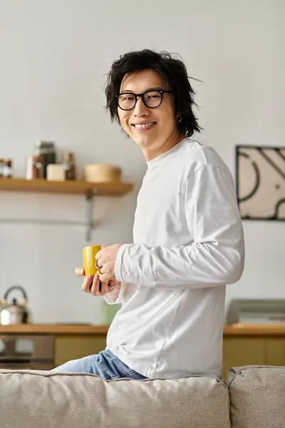 Joven de ojos brillantes sonríe mientras sostiene una taza amarilla en casa, exudando calidez y encanto. - foto de stock