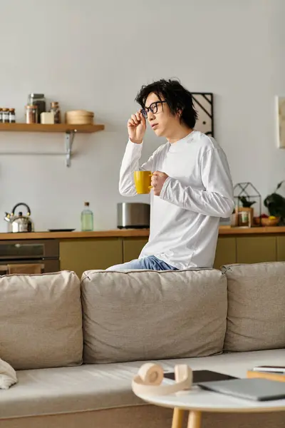 Un jeune homme avec des lunettes élégantes tient une tasse jaune tout en réfléchissant dans sa maison confortable. — Photo de stock