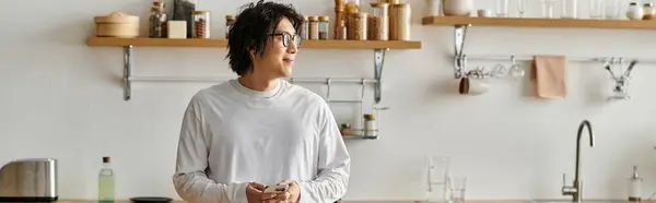 A stylish young asian man stands in a chic kitchen, casually holding his phone and smiling, banner — Stock Photo