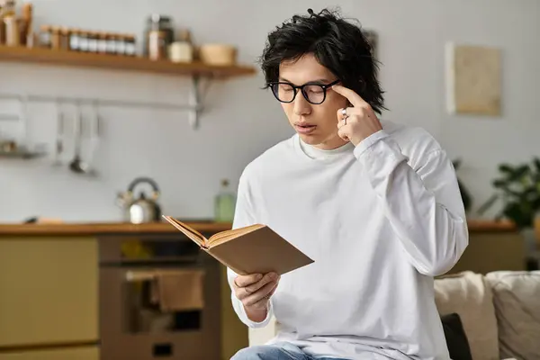 Ein hübscher junger asiatischer Mann sitzt gemütlich zu Hause und konzentriert sich mit einem Buch auf sein Studium. — Stockfoto