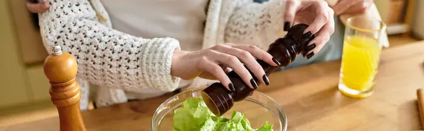 Young woman cooking salad and adding some pepper in it, next to her boyfriend. — Stock Photo