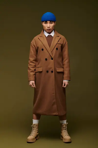Un joven, guapo hombre asiático con confianza modelos de un elegante abrigo marrón y gorro azul. - foto de stock