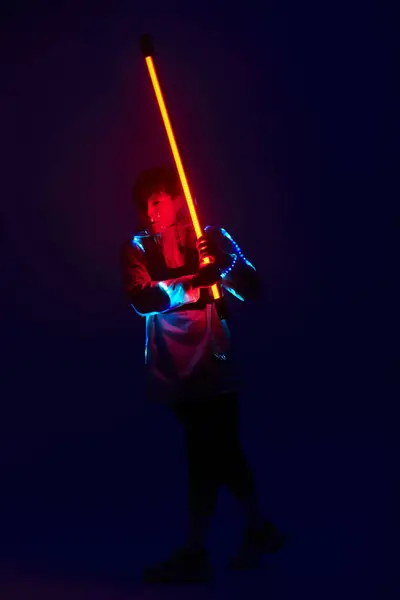 Un joven confiado con un atuendo moderno empuña una espada brillante bajo una iluminación dramática. - foto de stock