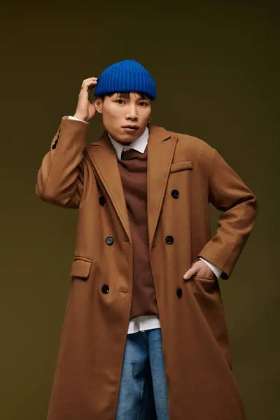 Un joven guapo hombre asiático posa con estilo en un abrigo marrón rico y sombrero azul, exudando confianza. — Stock Photo