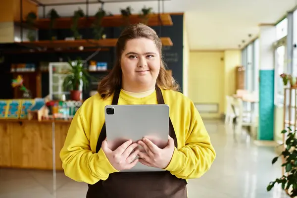 A strong young woman with Down syndrome stands in a colorful cafe, holding a tablet confidently. — Stock Photo