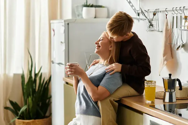 Un ragazzo abbraccia sua madre mentre lei si gode un drink caldo nella loro luminosa cucina, mostrando il loro legame. — Foto stock