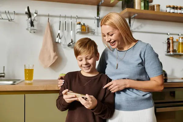 Una donna e il suo giovane figlio sorridono insieme mentre utilizzano uno smartphone nella loro cucina moderna. — Stock Photo