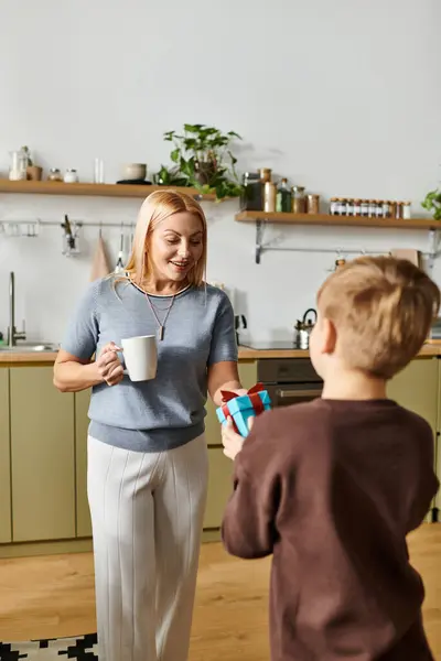 A mother smiles as her son gives her a colorful gift in their bright kitchen. — Stock Photo