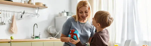 Mother smiles warmly as her son excitedly presents a wrapped gift in their bright kitchen setting — Stock Photo
