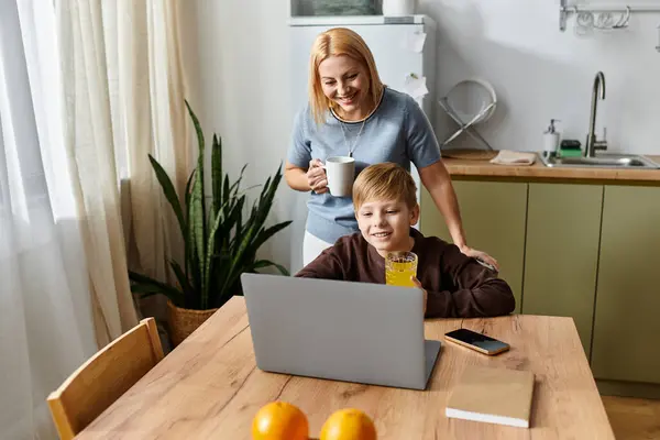 A joyful mother smiles at her son while he enjoys a refreshing drink and interacts with a laptop. — Stock Photo