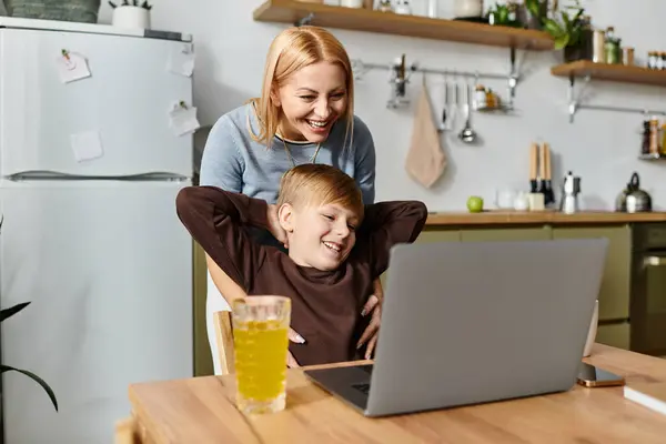 A joyful mother supports her son while they engage with a laptop in their cozy kitchen. — Stock Photo
