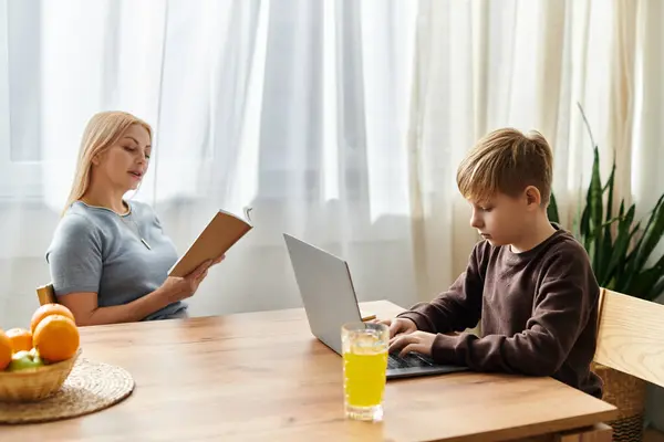 A mother reads a book while her son works on his laptop at a sunlit table of fruits. — Stock Photo