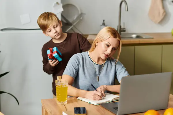A mother focuses on her writing as her son surprises her with a small gift in their cozy kitchen. — Stock Photo