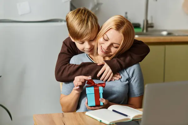Una madre y un hijo disfrutan de un momento conmovedor mientras desenvuelven un regalo en su acogedora cocina. - foto de stock