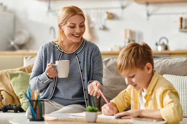 A mother enjoys her drink while warmly supporting her son with his drawing. — Stock Photo
