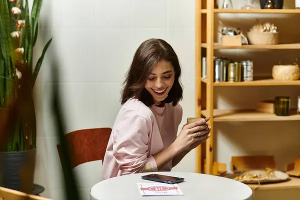 A delightful woman sits at a cafe table, sipping her drink while smiling at her phone. — Stock Photo