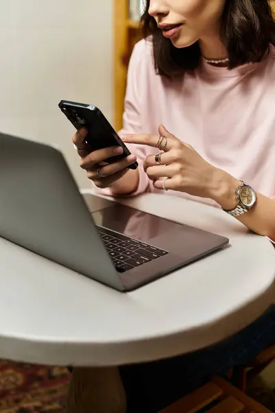 Молодая женщина фокусируется на ноутбуке, используя смартфон в современном кафе. — стоковое фото