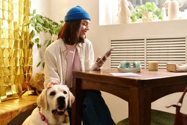 A young woman smiles at her phone while her labrador sits beside her in a cozy cafe. — Stock Photo