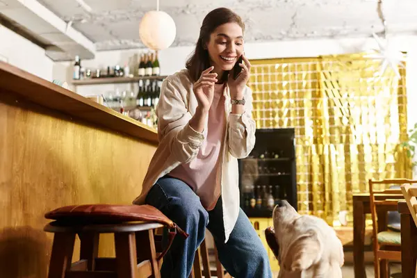 Una joven disfruta de una llamada telefónica mientras su labrador se acerca juguetonamente a ella en un café. — Stock Photo