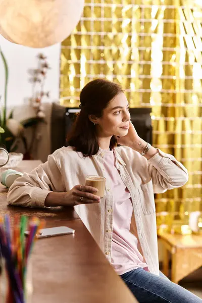 A pretty young woman relaxes at a modern cafe, sipping her drink and lost in thought. — Stock Photo