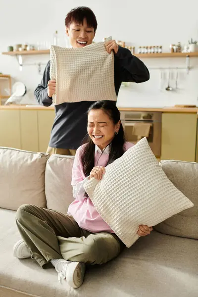 A joyful young asian couple engages in a lighthearted pillow fight at home, exuding happiness. — Stock Photo