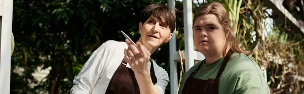 In a lush greenhouse, a mature woman helps her young colleague with Down syndrome. — Stock Photo