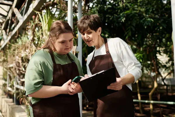 In a bright greenhouse, a mature woman works with her young colleague with Down syndrome. — Stock Photo