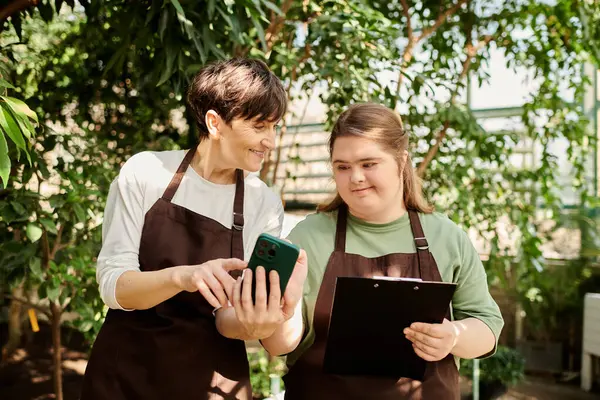 A mature woman guides her young colleague with Down syndrome while working in a lush greenhouse. — Stock Photo