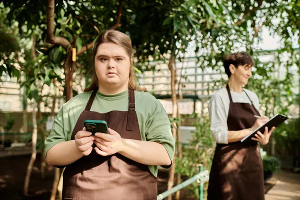 A mature woman watches her young colleague with Down syndrome working in a lush greenhouse. — Stock Photo