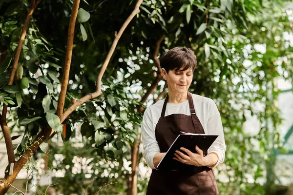 Mujer madura con el pelo corto observa las plantas y toma notas en un invernadero vibrante. - foto de stock