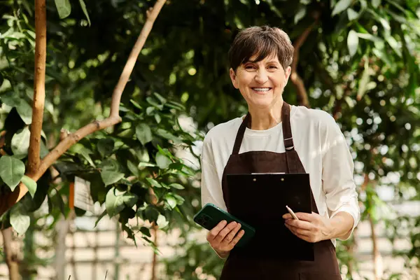 Donna matura si trova orgogliosamente in una serra, sorridendo mentre gestisce i suoi compiti vegetali con facilità. — Foto stock