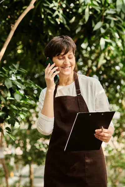 A confident mature woman with short hair is engaged in work while using a phone amidst greenery. — Stock Photo
