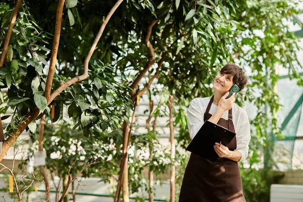 Mature woman enjoys her work in a vibrant greenhouse, connecting with nature and clients. — Stock Photo