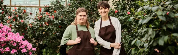 A mature woman assists her young colleague with Down syndrome in a lively greenhouse. — Stock Photo