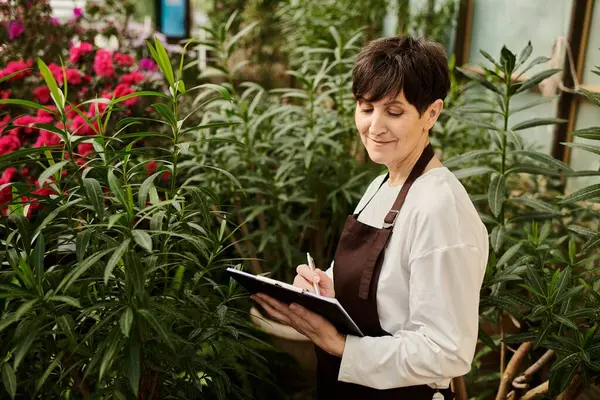 Mature woman working effortlessly in a greenhouse, taking notes and enjoying the vibrant flora. — Stock Photo