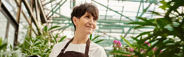 Mature woman works proudly in a thriving greenhouse, engaging with beautiful plants around her. — Stock Photo