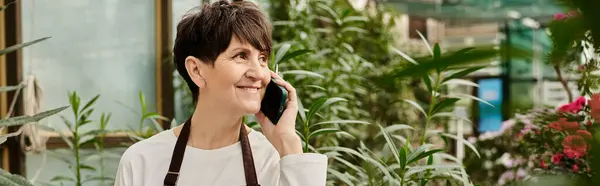 Mature woman wearing an apron happily talks on her phone amidst the lush greenery of the greenhouse — Stock Photo