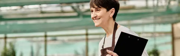 Donna matura porta una cartellina mentre lavora gioiosamente in una lussureggiante serra piena di verde. — Foto stock