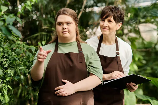 Em uma estufa exuberante, uma mulher ajuda seu jovem colega com síndrome de Down a cuidar de plantas. — Fotografia de Stock