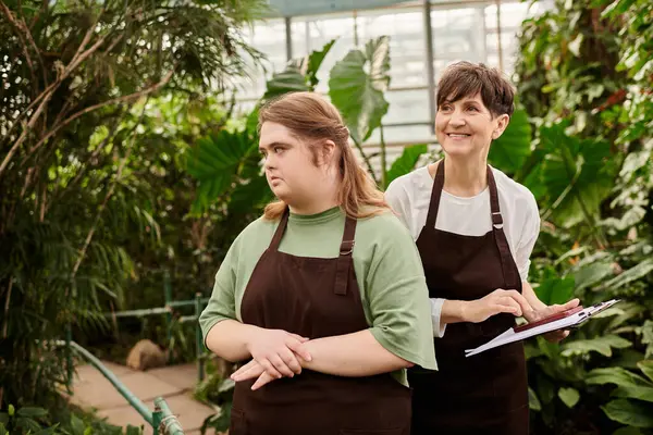 In a lush greenhouse, a mature woman assists her young colleague with Down syndrome. — Stock Photo
