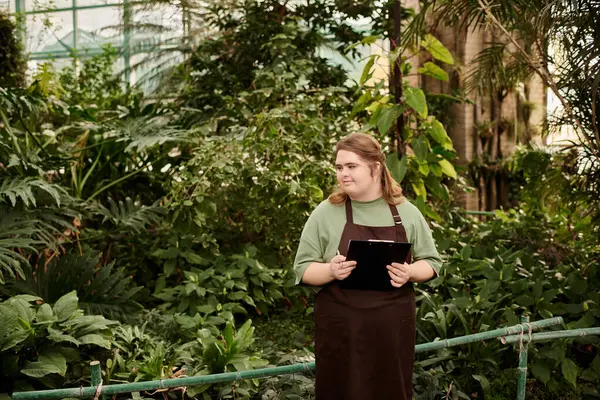In a vibrant greenhouse, a young woman with Down syndrome confidently tends to lush plants. — Stock Photo